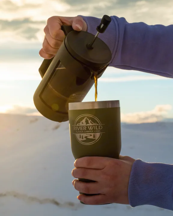 A person pours coffee into a River Wild Alaska Camper Vans mug amidst snowy mountains, perfect for an RV rental journey.. Dodge Promaster 2500 (Class B) 2022
