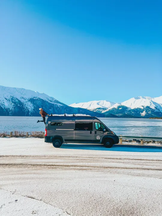 Explore winter in an **RV rental**! Gray van on snowy road, person hanging out of the back door, beside a frozen lake & mountains.. Dodge Ram 2500 (Class B) 2021