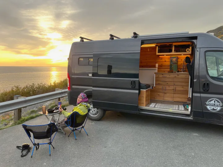 A person relaxes outside a gray RV rental van, dining at sunset by the ocean, its custom wooden interior visible.. Dodge Ram 2500 (Class B) 2021