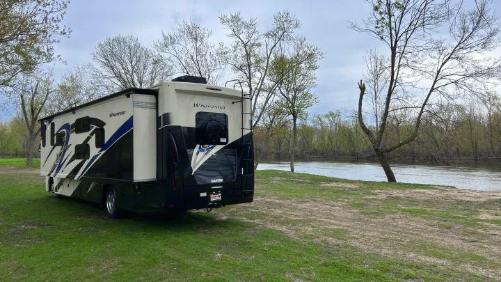 Riverside serenity: A large Windsport RV rental parked on a grassy bank by a calm river, surrounded by trees.. Thor Windsport 2023