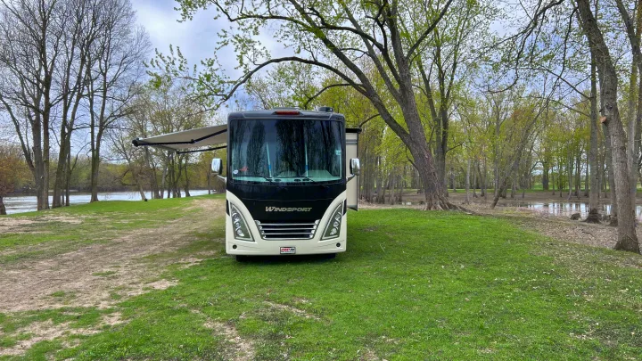 Scenic RV rental: a black and white motorhome with an extended awning by a river and trees in a natural park.. Thor Windsport 2023