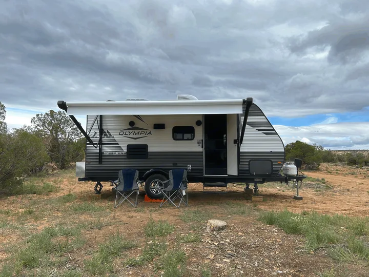 An RV rental, an Olympia Sport travel trailer, with its awning extended and chairs out, parked in a dry, natural camp.. Olympia Sport 2022