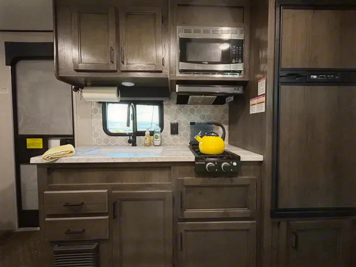 RV rental kitchen interior with dark cabinets, white counters, sink, microwave, stove with a yellow kettle, and refrigerator.. Olympia Sport 2022