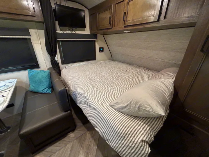 Inside an RV rental, a compact bedroom features a striped bed, overhead cabinets, and a small dinette.. Olympia Sport 2022