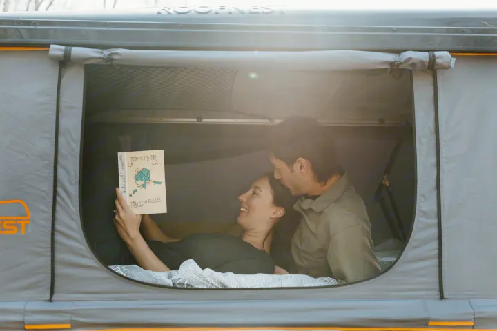 A happy couple reading "Travels in Alaska" by John Muir inside a Roofnest rooftop tent, cozy for an RV rental camping trip.. Jeep Wrangler 2023