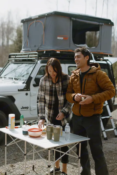 Campers prep food on a portable table beside their Jeep and rooftop tent, an ideal setup for a compact **RV rental** trip.. Jeep Wrangler 2023