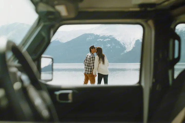 Cozy RV rental view: Couple shares a kiss by a serene lake and snow-capped mountains, framed by the vehicle interior.. Jeep Wrangler 2023