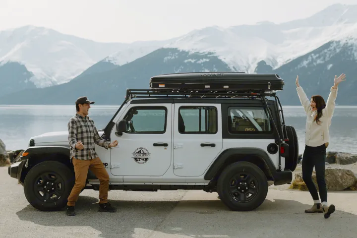 Excited travelers with their white RV rental Jeep, featuring a Roofnest rooftop tent, from River Wild Alaska, by a scenic lake.. Jeep Wrangler 2023