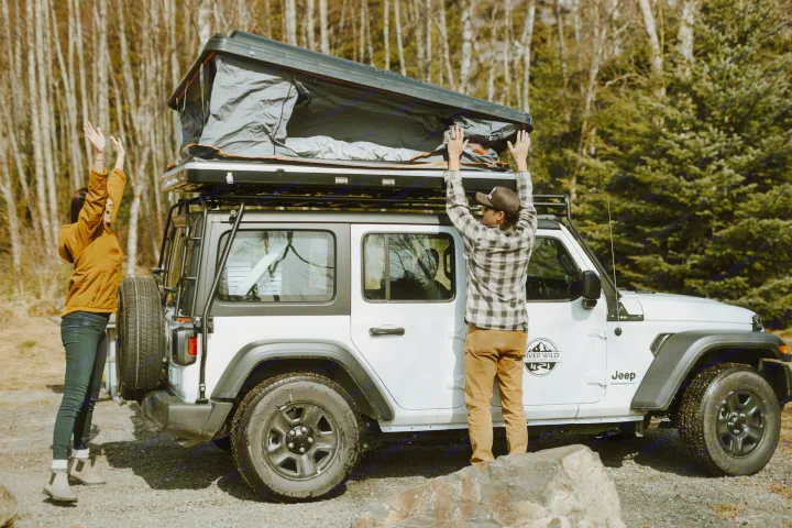 Two people open a rooftop tent on a white Jeep Wrangler RV rental for a camping adventure.. Jeep Wrangler 2023