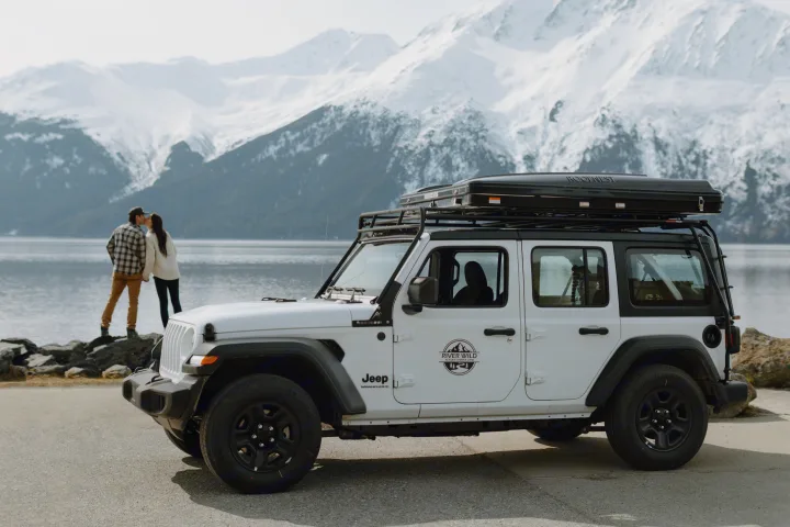 Scenic Alaska RV rental: White Jeep Wrangler with rooftop tent & a couple by a lake with snowy mountains.. Jeep Wrangler 2023