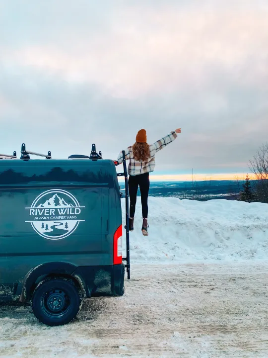 A woman on an Alaska RV rental van's ladder points to a vast, snowy landscape under a colorful sky.. Dodge Ram 2500 (Class B) 2021