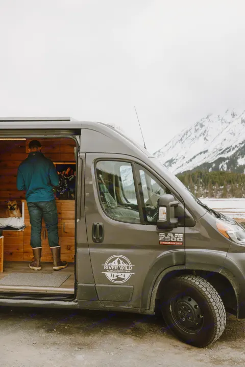 A custom RV rental van, River Wild Alaska Camper Vans, with its side door open revealing a person and dog inside, surrounded by snowy mountains.. Dodge Ram 2500 (Class B) 2021