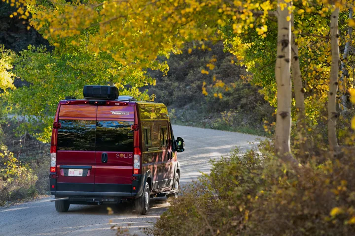 Red Winnebago Solis rv rental driving on a scenic autumn road, surrounded by vibrant yellow fall foliage.. Winnebago Solis PX 2023