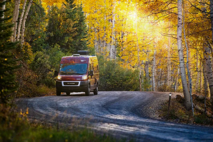 Experience autumn beauty! A maroon RV rental camper van drives a scenic gravel road through brilliant yellow aspen trees and sunlight.. Winnebago Solis PX 2023