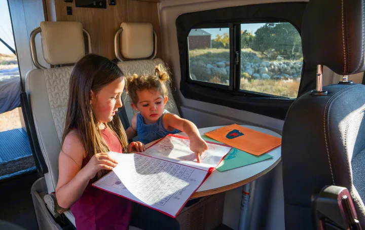 Two girls learn math and shapes in their RV rental. They sit at a table inside the camper van, with nature visible outside the window.. Winnebago Solis PX 2023