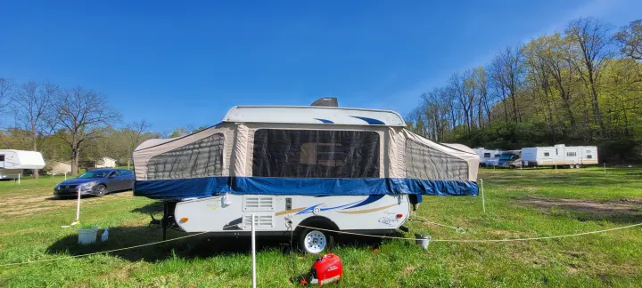 An RV rental pop-up camper is set up in a grassy campground, with other RVs visible in the background under a clear blue sky.. Coleman clipper 2011