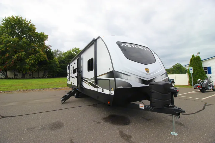 White Astoria travel trailer, ideal for an RV rental, parked on asphalt with steps extended. Trees and a building are in the background.. Dutchmen Astoria 2023