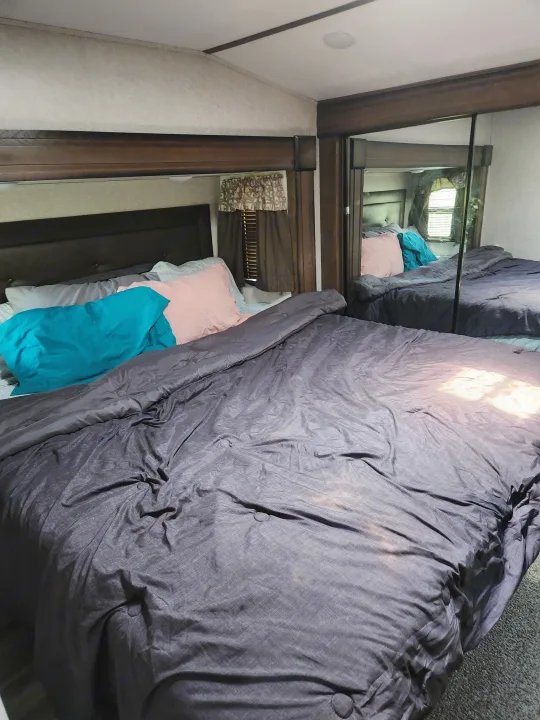 Cozy RV rental bedroom with dark bedding, pink pillow, and mirrored closet reflecting a window view.. Keystone RV Cougar 2019