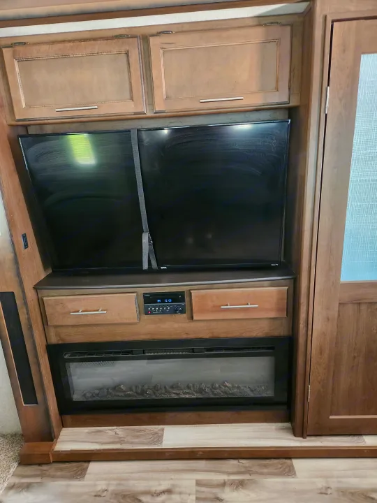 Inside an RV rental: entertainment setup with a secured TV, media control, electric fireplace, and wood cabinets.. Keystone RV Cougar 2019
