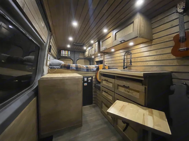 This RV rental showcases a warm wood-paneled interior with a built-in kitchen, bed, and cozy lighting.. Mercedes-Benz Sprinter 2020
