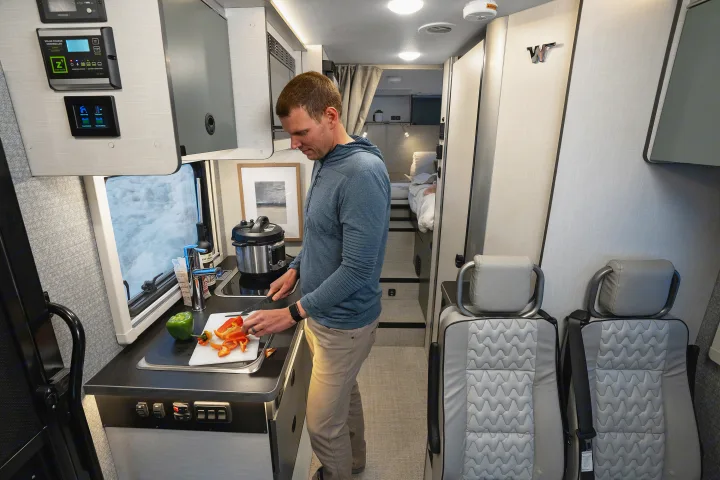 An RV rental features a compact kitchen where a man prepares food, with controls and a cozy bed area visible.. Winnebago EKKO POP TOP 2027