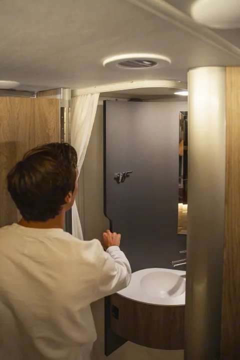 Compact RV rental bathroom: A man adjusts a grey panel next to the white sink and shower curtain.. Winnebago EKKO POP TOP 2027