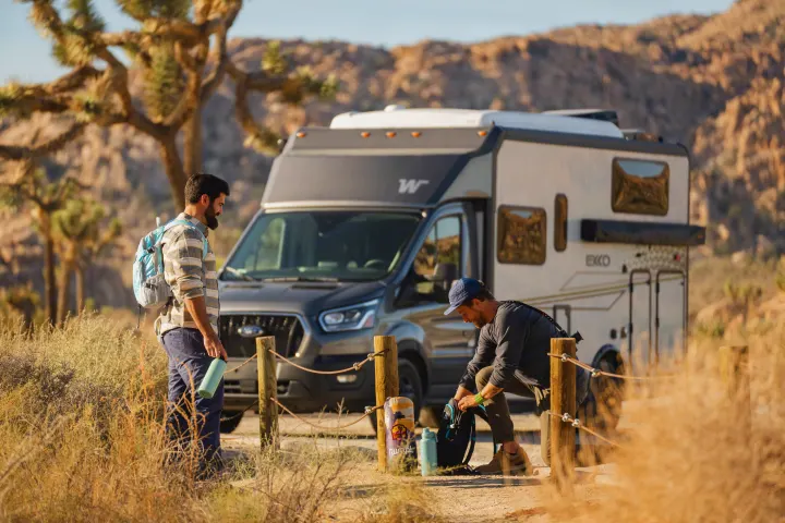Two men with backpacks and water bottles are next to their RV rental at a sunny desert campsite, preparing for an outing.. Winnebago EKKO POP TOP 2027