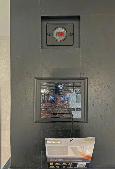 Inside an RV rental: a control panel featuring a 12.2V DC meter and a DRIVEN entertainment system showing AM 12:06.. Forest River Cherokee Wolf Pup Black Label 2022