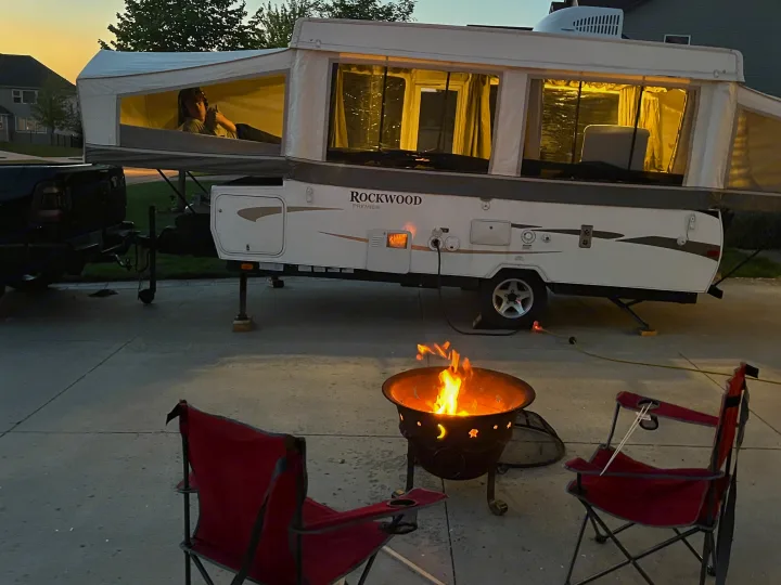 Evening scene: a glowing pop-up rv rental camper with a person inside, next to a warm campfire and red chairs.. Forest River Rockwood Premier 2009