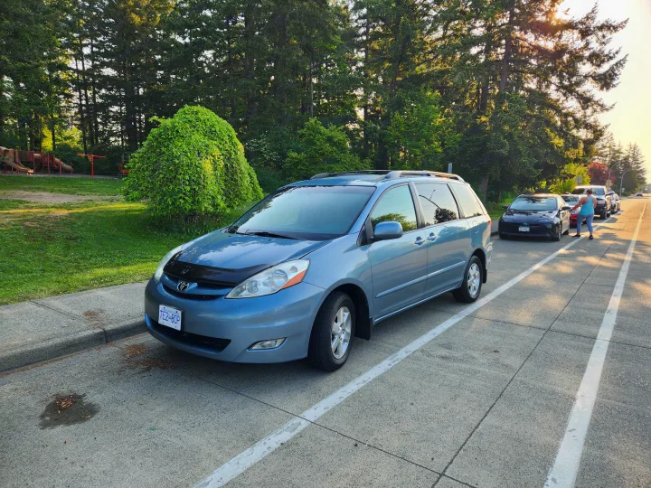 A light blue minivan parked curbside by a sunny park with tall trees. A practical choice for travel, even without an rv rental.. Toyota Sienna 2008
