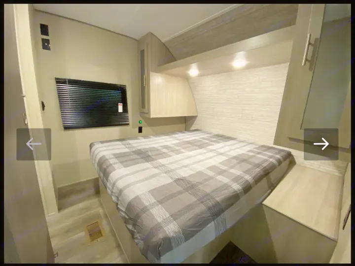 This RV rental bedroom features a comfy bed with grey plaid bedding, overhead cabinets, and a window with blinds.. Coachmen Catalina 2023