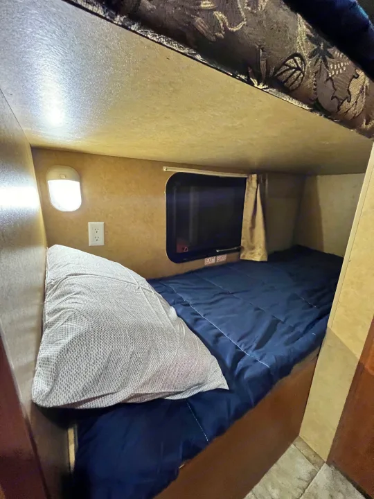 Inside an RV rental, showing a lower bunk bed with blue bedding, a pillow, a wall light, and a window.. Jayco Jay Flight Swift 154 BH Slx 2013