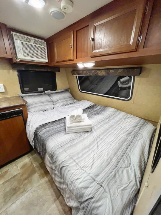 This cozy RV rental interior features a neatly made bed with striped bedding, side windows, an AC unit, and overhead wooden storage.. Jayco Jay Flight Swift 154 BH Slx 2013