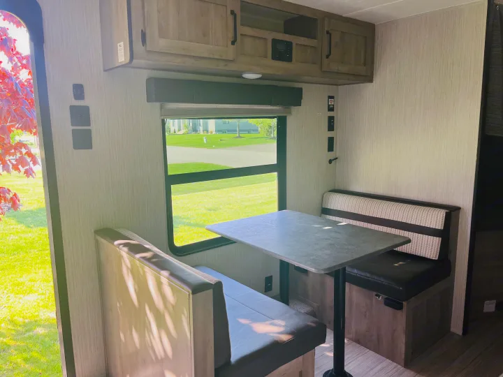 This inviting RV rental interior showcases a cozy dinette with a table, two benches, and a large window offering a sunny green view.. Palomino PUMA 22RBC 2022