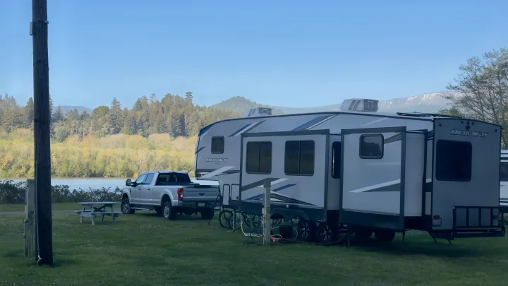 Spacious RV rental and truck at a scenic riverside campsite with lush trees and snowy mountains.. Forest River Cherokee Arctic Wolf 2022