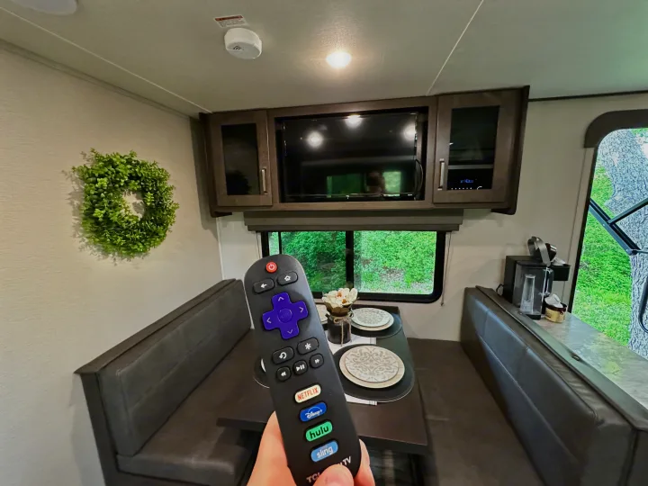 RV rental interior: A hand holds a Roku remote, ready to control the TV in the cozy dinette area. Perfect camper entertainment.. Grand Design Transcend Xplor 2022