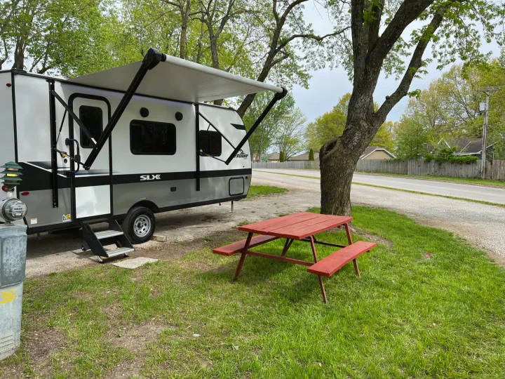 An RV rental is set up at a grassy site with an extended awning and red picnic table. Steps lead into the camper.. Jayco Jay Flight SLX 2022