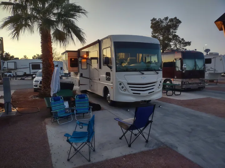 An RV campground at sunset featuring a white Class A motorhome, camping chairs, a palm tree, and other rv rental units.. Flair-Fleetwood Flair-Fleetwood Motorhome 2020