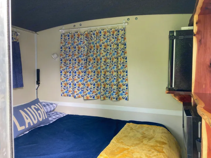 Cozy RV rental interior featuring a comfortable bed with blue bedding, floral curtains, and a compact kitchenette.. Runaway Rouser 2023