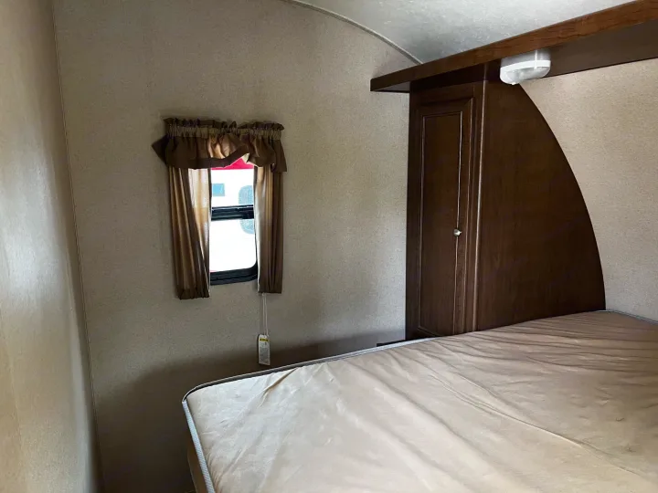 Cozy RV rental bedroom showing a comfortable mattress, window with curtains, and dark wood storage cabinet.. Shasta Oasis 2016