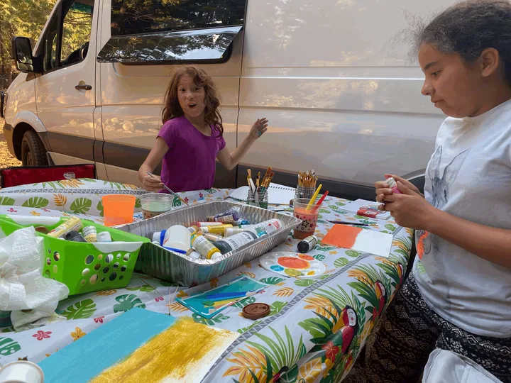 Two girls enjoy outdoor painting near their RV rental, surrounded by vibrant art supplies, with one girl showing surprise.. Mercedes-Benz Sprinter 2017