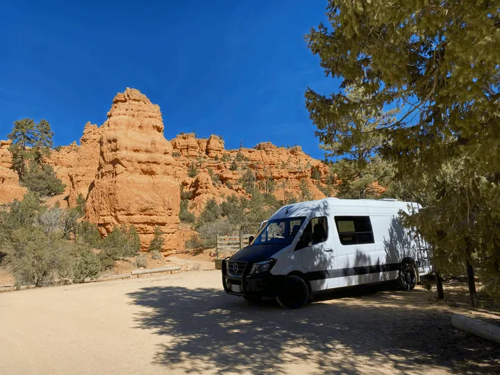 A white Mercedes Sprinter RV rental parked on a dirt road, framed by towering red rock formations and a bright blue sky.. Mercedes-Benz Sprinter 2017