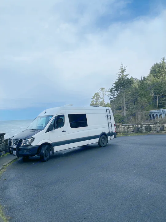 White RV rental on a scenic coastal road. This camper van is parked by ocean waves, a stone wall, an arched bridge, and trees.. Mercedes-Benz Sprinter 2017