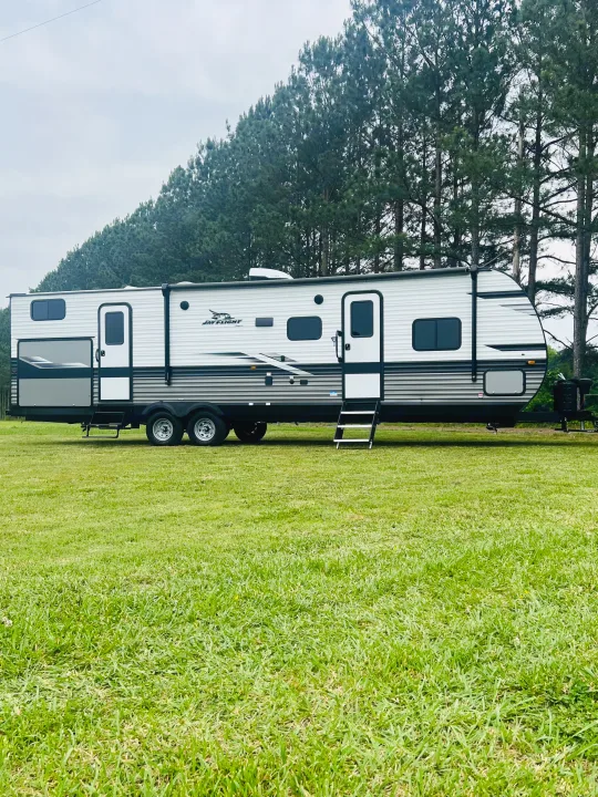 A white and grey Jay Flight travel trailer for RV rental, parked on green grass. Tall pine trees line the background.. Jayco Jayflight 324BDS 2023