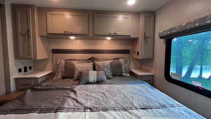 Modern RV rental bedroom with a gray bed, multiple storage cabinets above and beside, integrated nightstands, and a large window.. Thor Windsport 2023