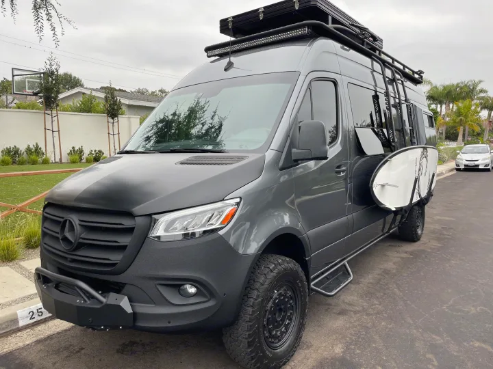 Customized gray Mercedes Sprinter van with off-road tires, roof rack, and surfboard, ready for adventure as an RV rental.. Mercedes-Benz Sprinter 2020