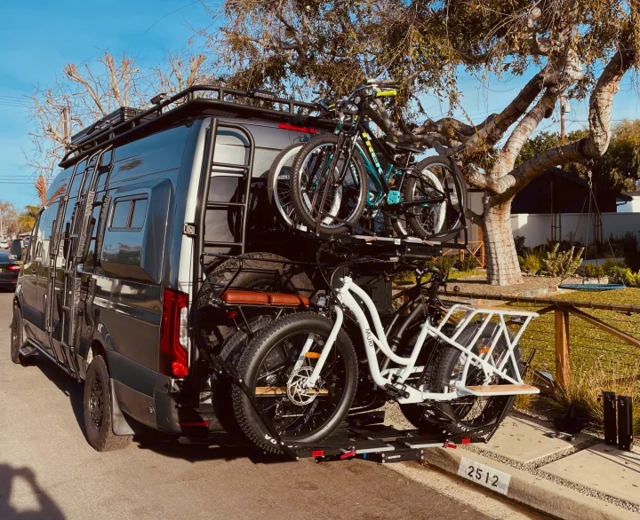 Adventure-ready RV rental van with roof rack, ladder, & a rear rack holding 4 bikes & spare tires, parked on a sunny street.. Mercedes-Benz Sprinter 2020