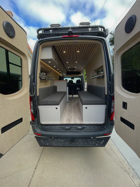 Spacious custom RV rental van interior, seen from rear with open doors, showing benches, wood ceiling, and roof rack.. Mercedes-Benz Sprinter 2020