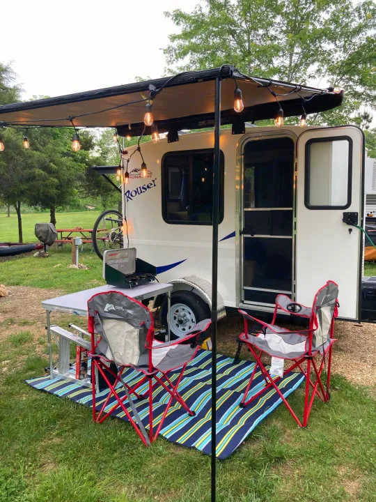 RV rental campsite: Compact trailer with string lights, awning, outdoor table, chairs & portable stove for cozy camping.. Runaway Rouser 2023