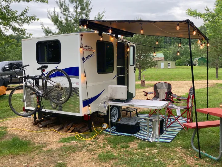 A small white rv rental camper, a Runaway Rouser, with a lighted awning, bike, and outdoor furniture for a cozy camp setup.. Runaway Rouser 2023